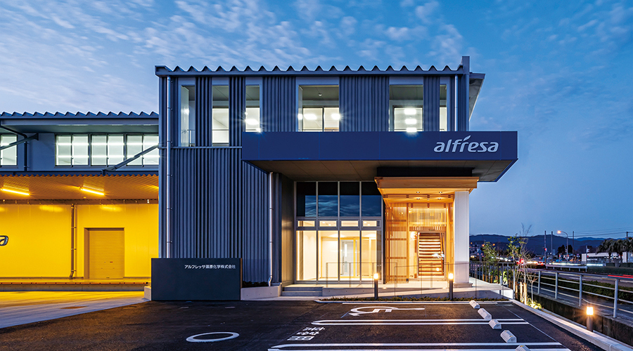 アルフレッサ篠原化学本社新社屋の夜景。入口や倉庫、窓がオレンジ色の明かりが灯されている。